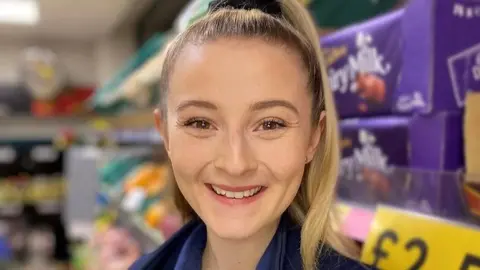 Hannah Lowther Smiling lady with blonde pony tail with dairy milk on supermarket shelves in background