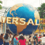 Crowd of people at an amusement park entrance with a large globe sculpture labeled "Universal" overhead, surrounded by colorful buildings and palm trees.